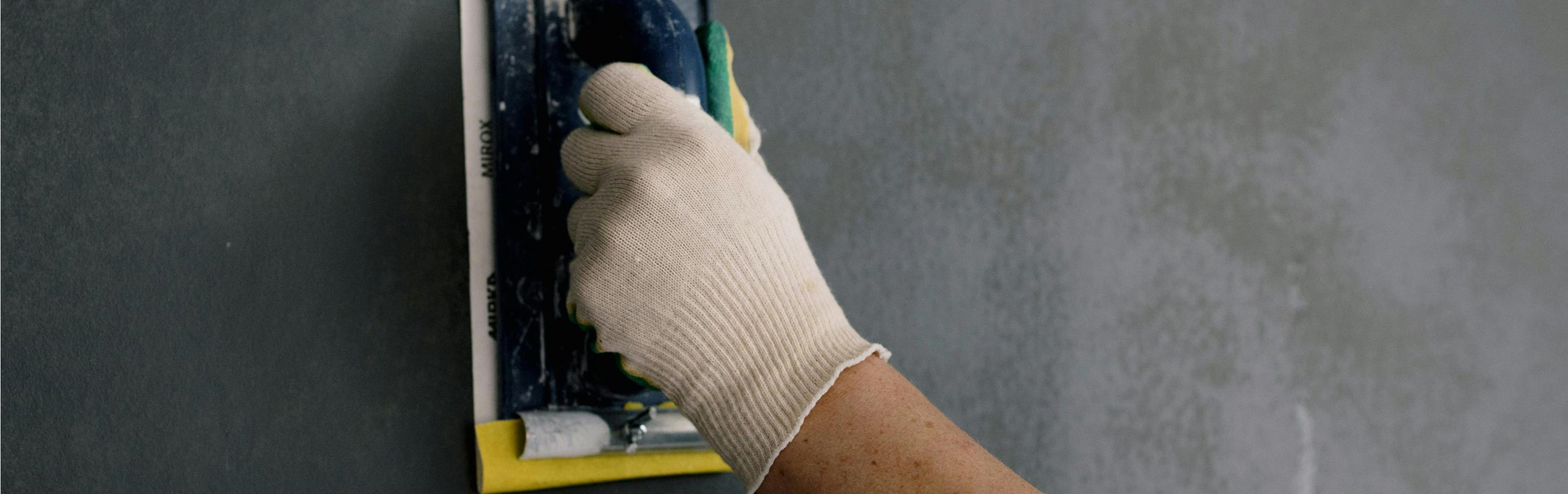 Hand mit Arbeitshandschuh glättet eine graue Wand mit einem Schleif- oder Glättwerkzeug.