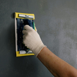 Hand mit Arbeitshandschuh glättet eine graue Wand mit einem Schleif- oder Glättwerkzeug.